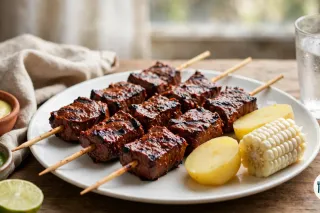 Tres palitos de anticuchos de corazón a la parrilla en un plato blanco con patata cocida y un trozo de choclo, junto a dos cuencos de salsa amarilla y verde y media lima